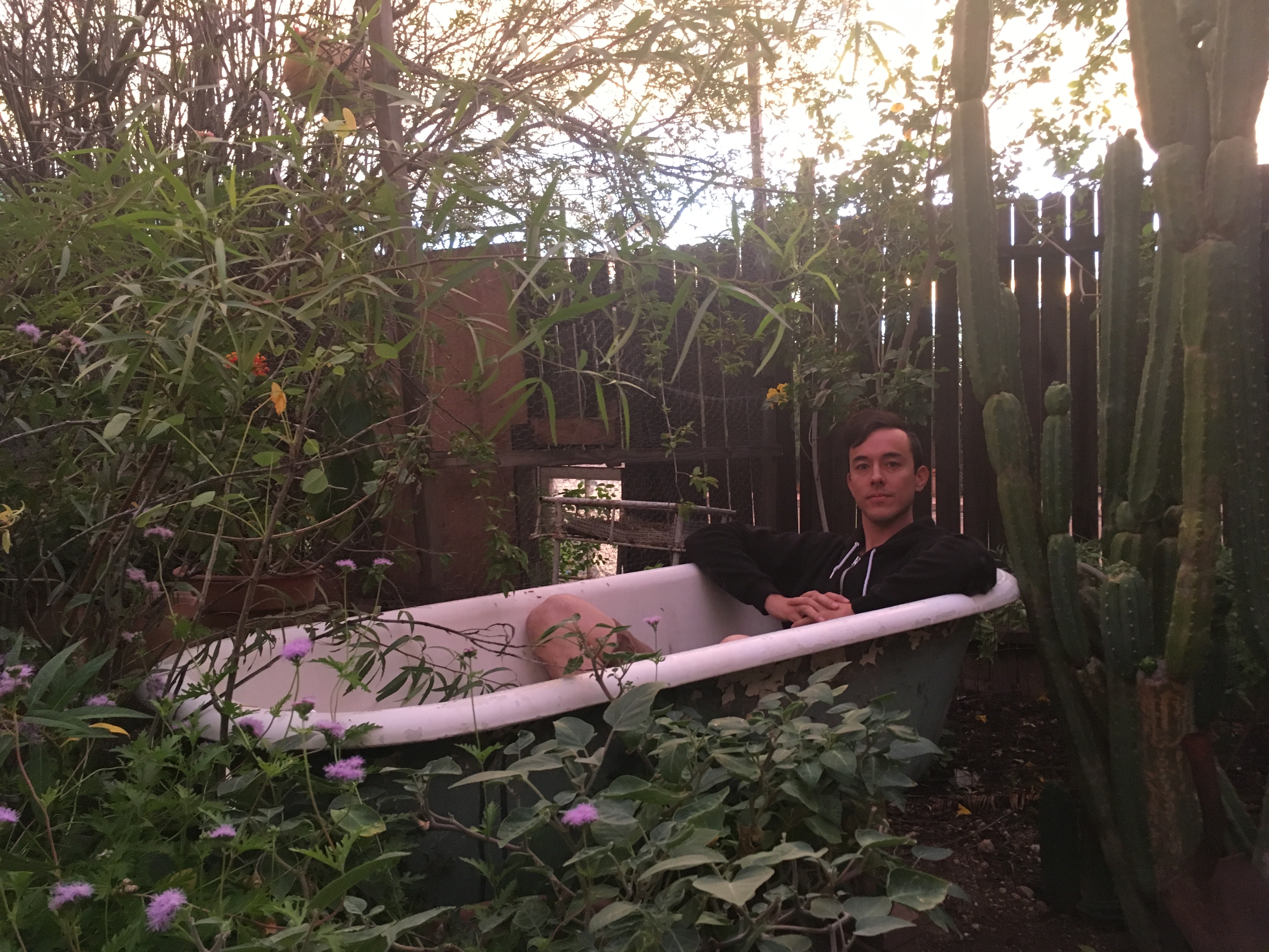 A man with black hair sits in a bathtub that has been placed in a backyard with plants growing all around.