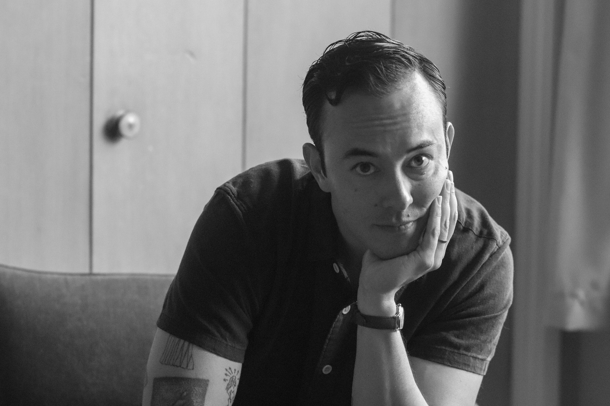 Black and white portrait of an Asian American man in his mid-30s. He wears a black shirt and has his hand cupping his chin.