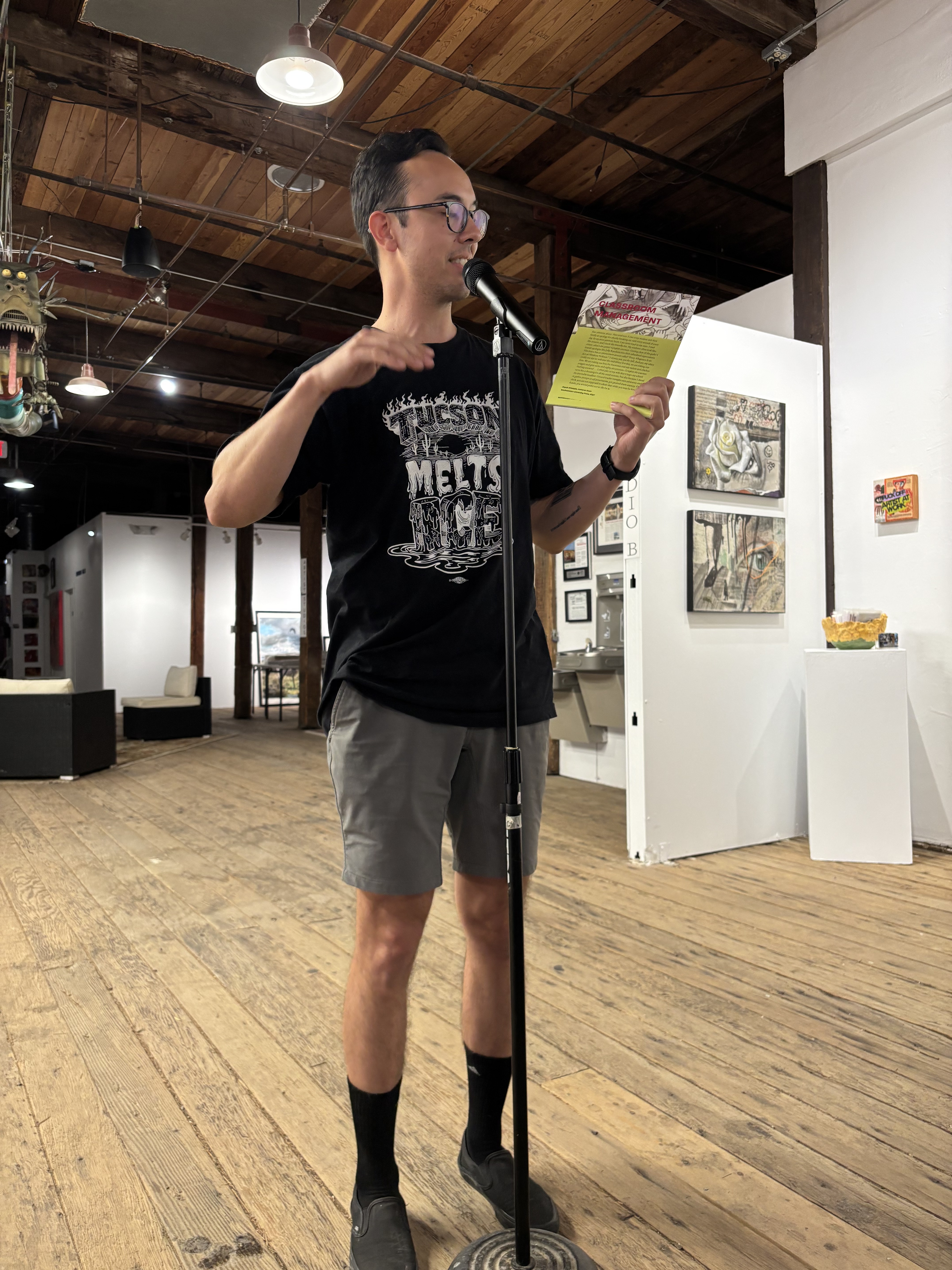 Man standing in front of mic, showing a poetry broadside to audience. He wears a black shirt that says "Tucson Melts ICE"